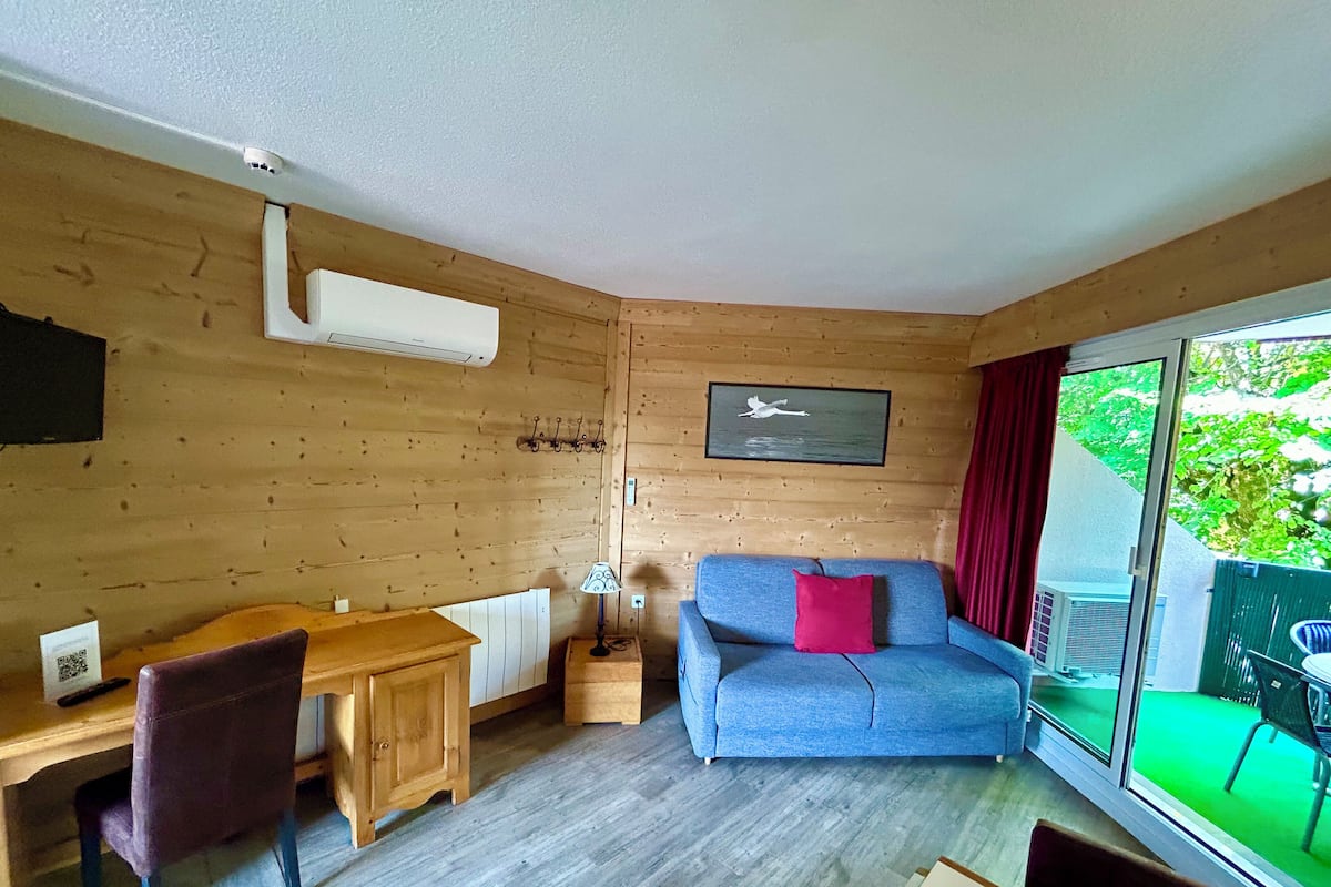 A cozy room with a wooden interior, a blue sofa with a red pillow, a wooden desk with a chair, and a large window offering a view of greenery.