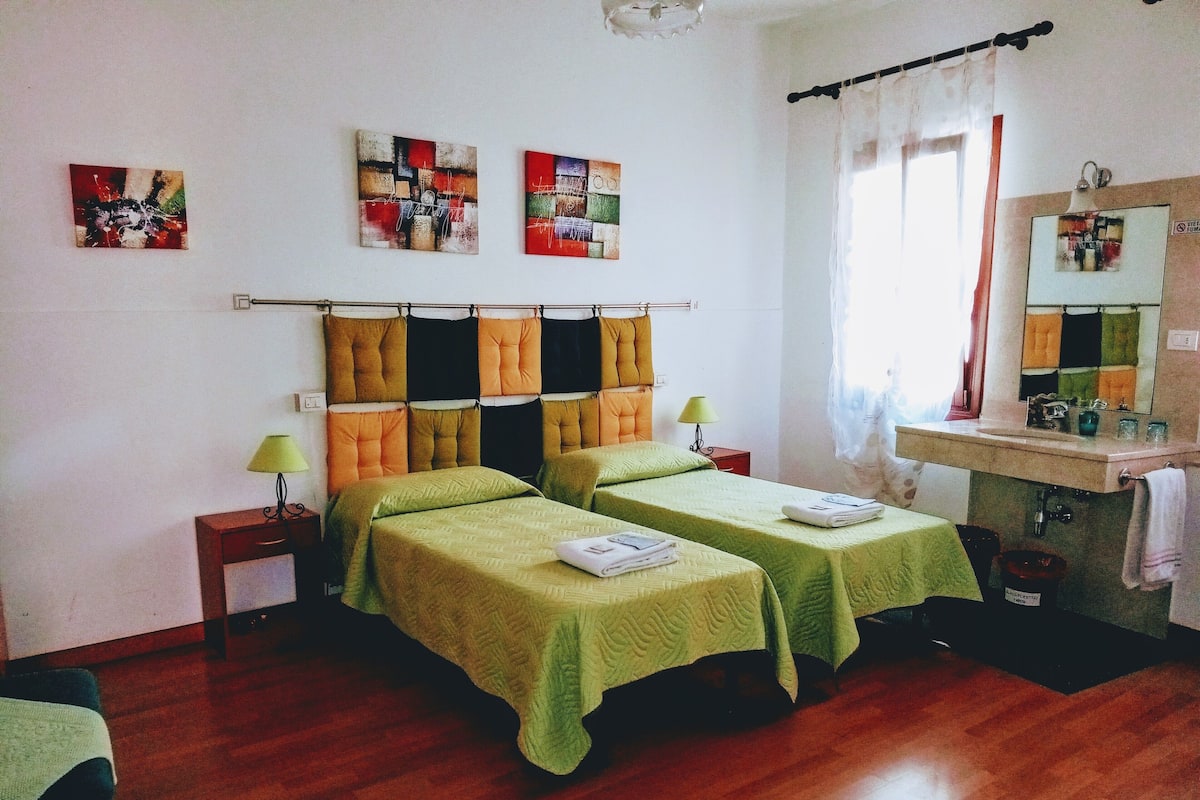 A hotel room with two beds, a small bedside table with a lamp, a bathroom with a sink and mirror, and artwork on the walls.