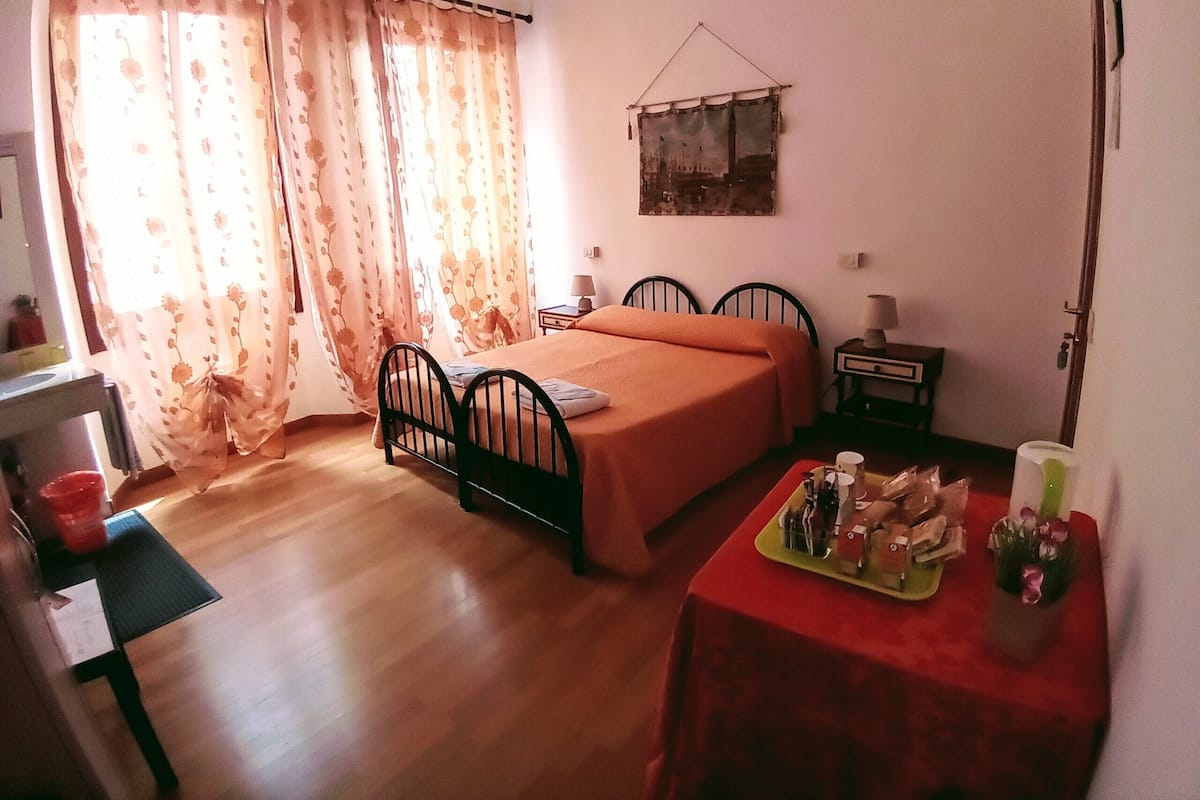 A bedroom with a bed, a desk, a chair, a small table with items, and a window with curtains.