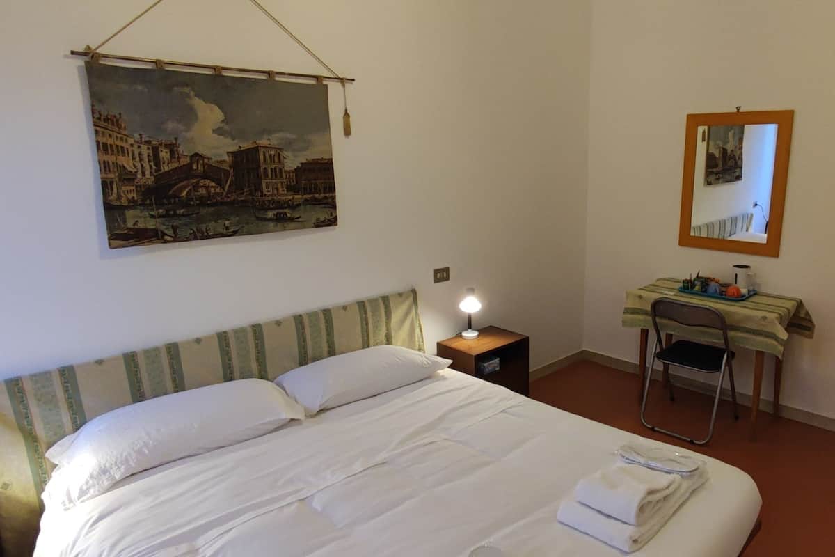 A hotel room with a bed, a desk with a chair, a nightstand, a lamp, a painting, and a mirror.
