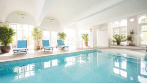 Una piscina interior con agua azul cristalina, rodeada de plantas en macetas y sillas reclinables azules.