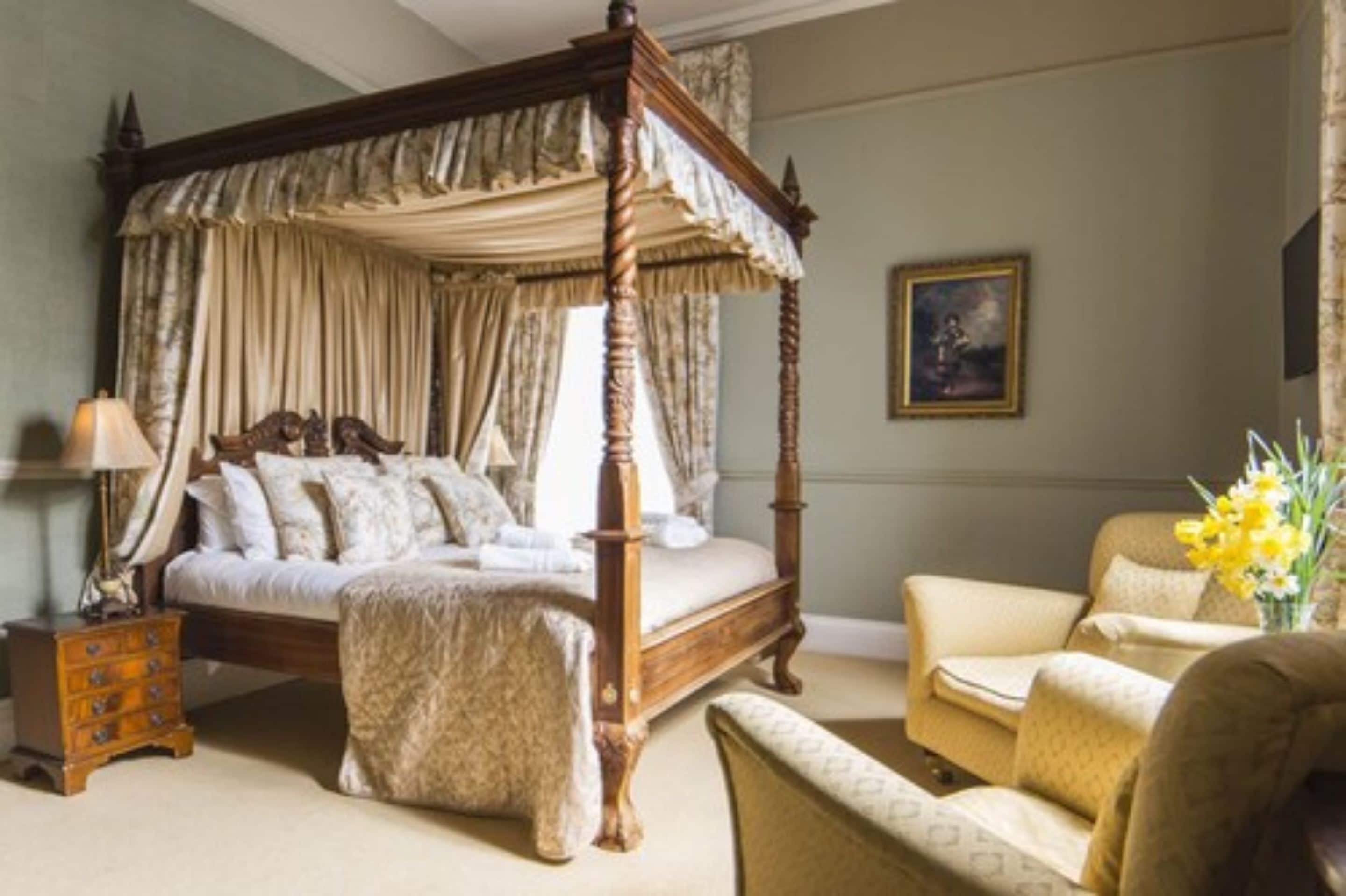 A four-poster canopy bed with a wooden frame and draped curtains, a bedside table with a lamp, a chair, and a vase with flowers.