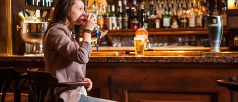 Una persona sentada en un bar, con diversas botellas y una grifería al fondo.