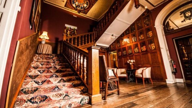 Una imponente escalera con barandillas ornamentales y alfombra estampada que conduce a un salón con paneles de madera y un candelabro.