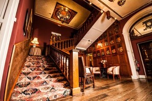 Un magnifique escalier, agrémenté de rampes décoratives et recouvert d’une moquette à motifs, mène à une pièce aux murs lambrissés et ornée d’un lustre.