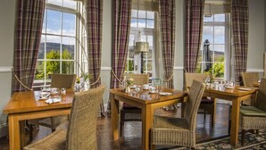 Un comedor con una mesa de madera y sillas de mimbre, ventanas amplias con cortinas a cuadros y vistas al exterior con vegetación.
