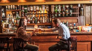 Dos personas sentadas en una barra, con una variedad de botellas de licor al fondo.