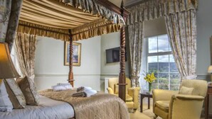 Una cama con dosel y cuatro columnas, con edredón acolchado, una mesita de noche con lámpara, una silla y vistas al exterior a través de una ventana.