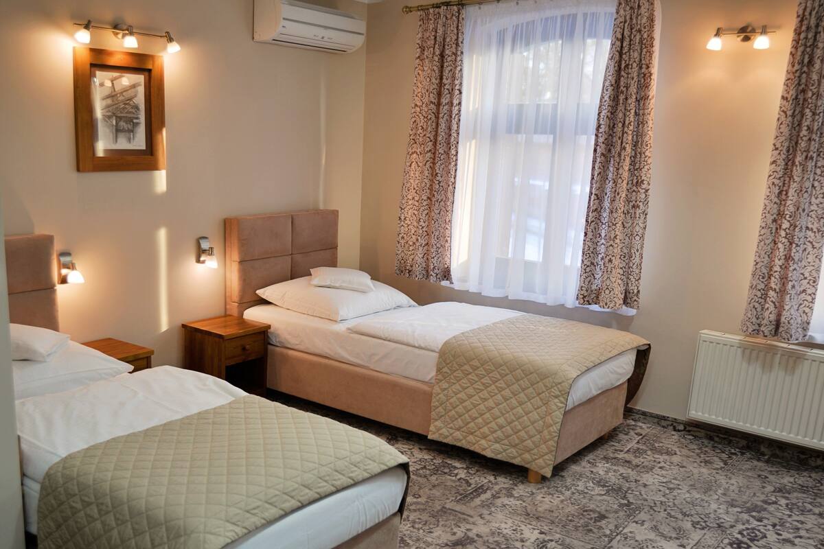 A hotel room with two beds, a large window with patterned curtains, a radiator, and wall-mounted lights.