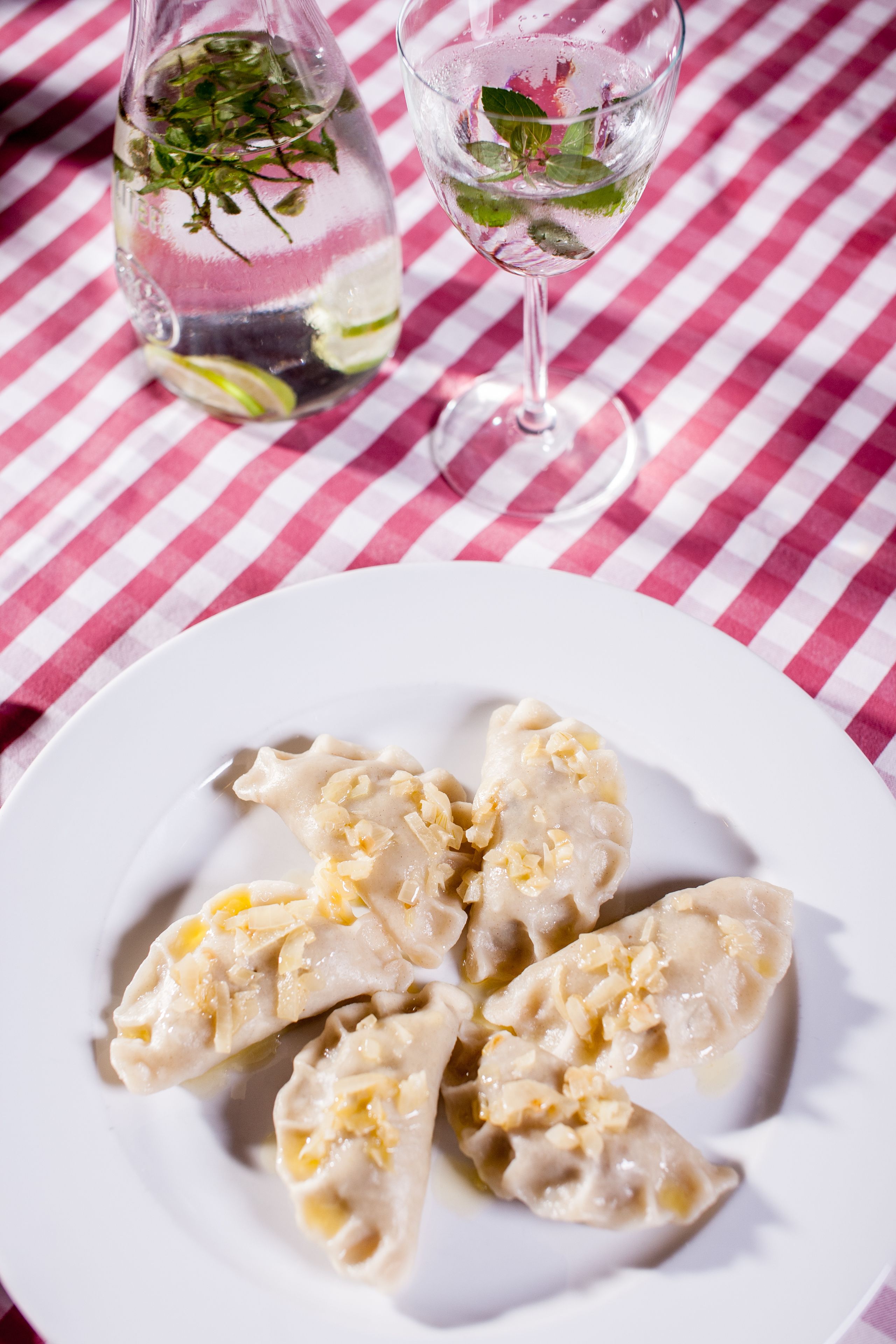 A plate of dumplings on a white plate, a glass of water, and a bottle of water with mint leaves on a checkered tablecloth.