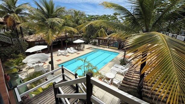 A pool area with lounge chairs and umbrellas, surrounded by palm trees and a building in the background.