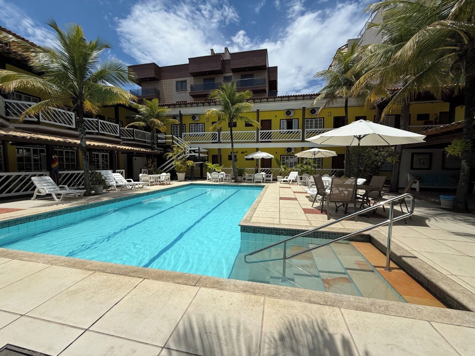 A swimming pool with lounge chairs and umbrellas, surrounded by palm trees and a multi-story building with balconies.