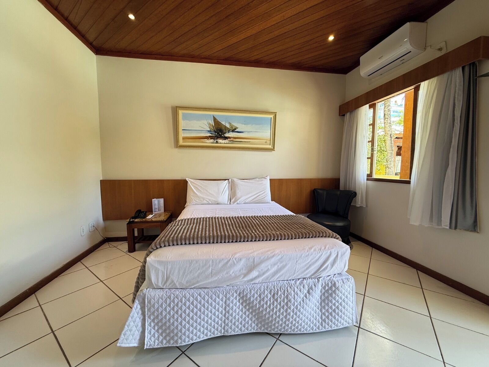 A hotel room with a bed, a nightstand, a chair, a painting on the wall, and a window with curtains.