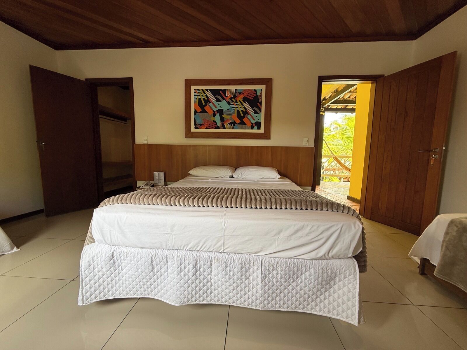 A bedroom with a large bed, a wooden ceiling, a painting on the wall, and a door leading to an outdoor area.