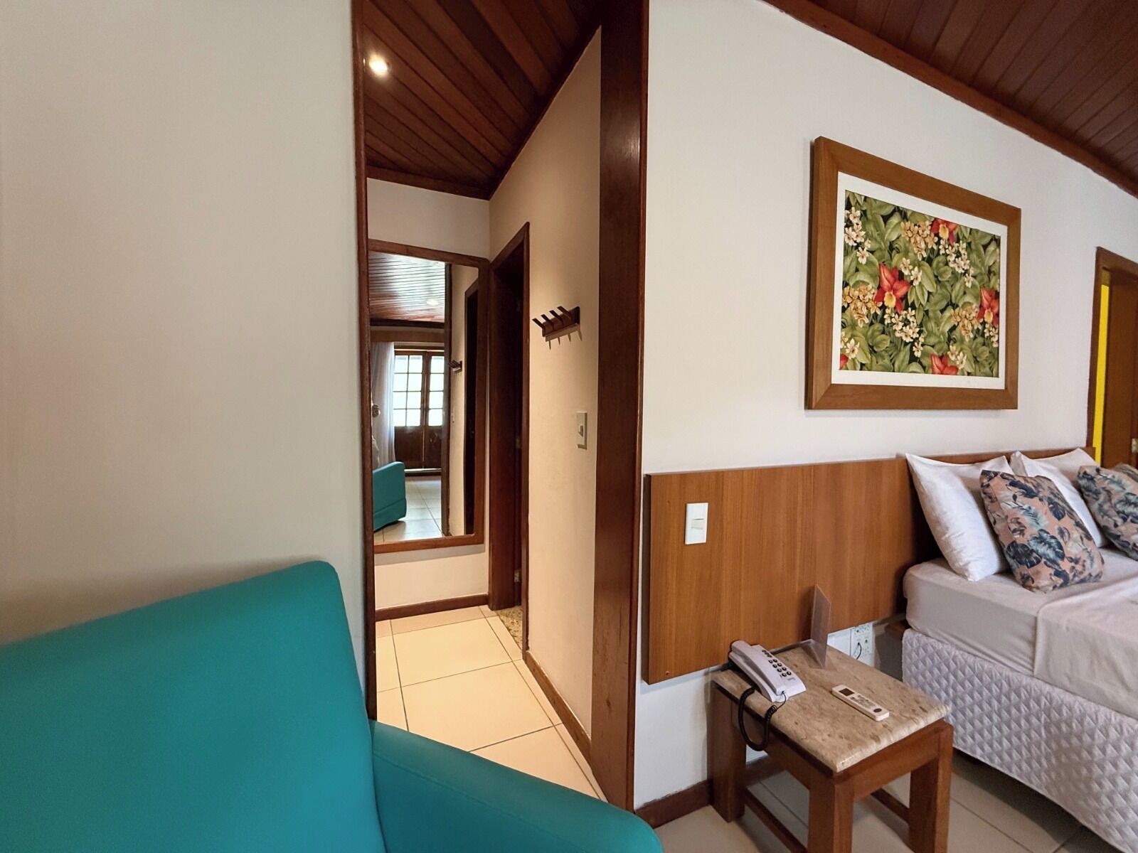 Une pièce avec un canapé de couleur turquoise, un lit agrémenté d’oreillers à motifs floraux, une tête de lit en bois et un tableau représentant des fleurs accroché au mur.