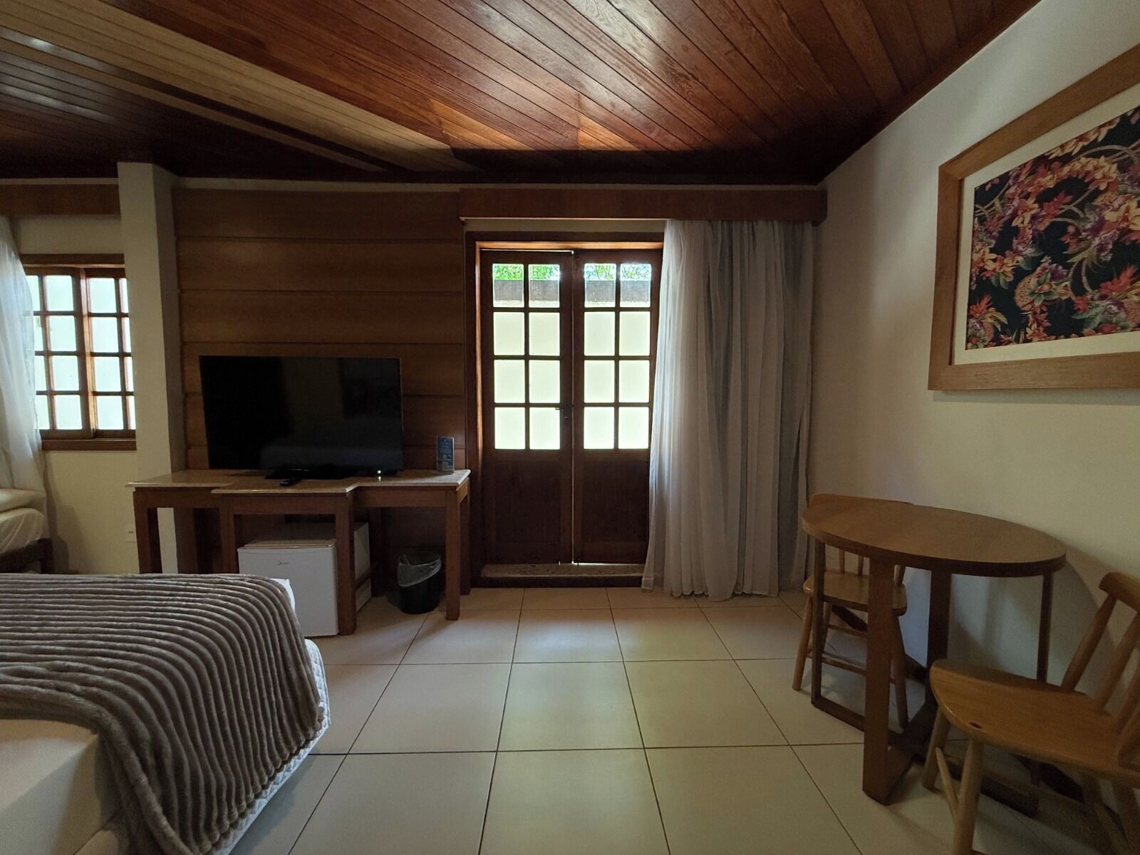 A cozy room with a wooden ceiling, a bed, a desk with a television, a window with curtains, and a painting on the wall.