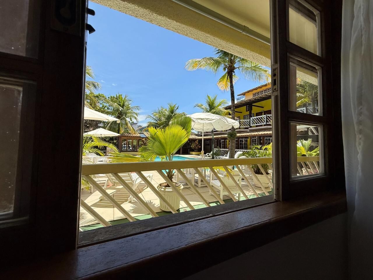 On aperçoit une piscine, des parasols et des chaises longues à travers une fenêtre ouverte.