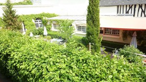 Un jardin luxuriant, doté d’un feuillage dense, un petit espace extérieur aménagé avec des sièges, et un bâtiment à la façade blanche et aux encadrements de fenêtres noirs.