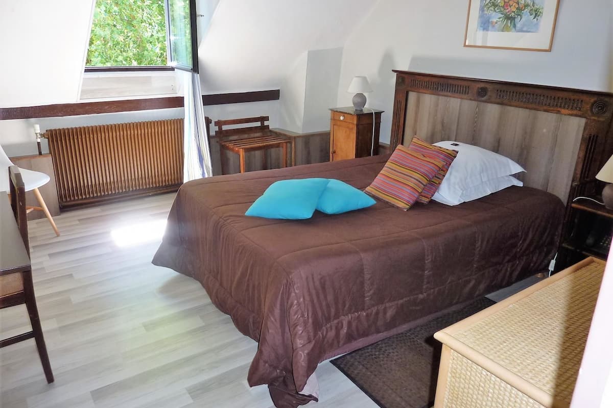 Una camera da letto con un letto grande, una testiera in legno, un comodino, una sedia, un termosifone e una finestra con vista su un paesaggio verde.