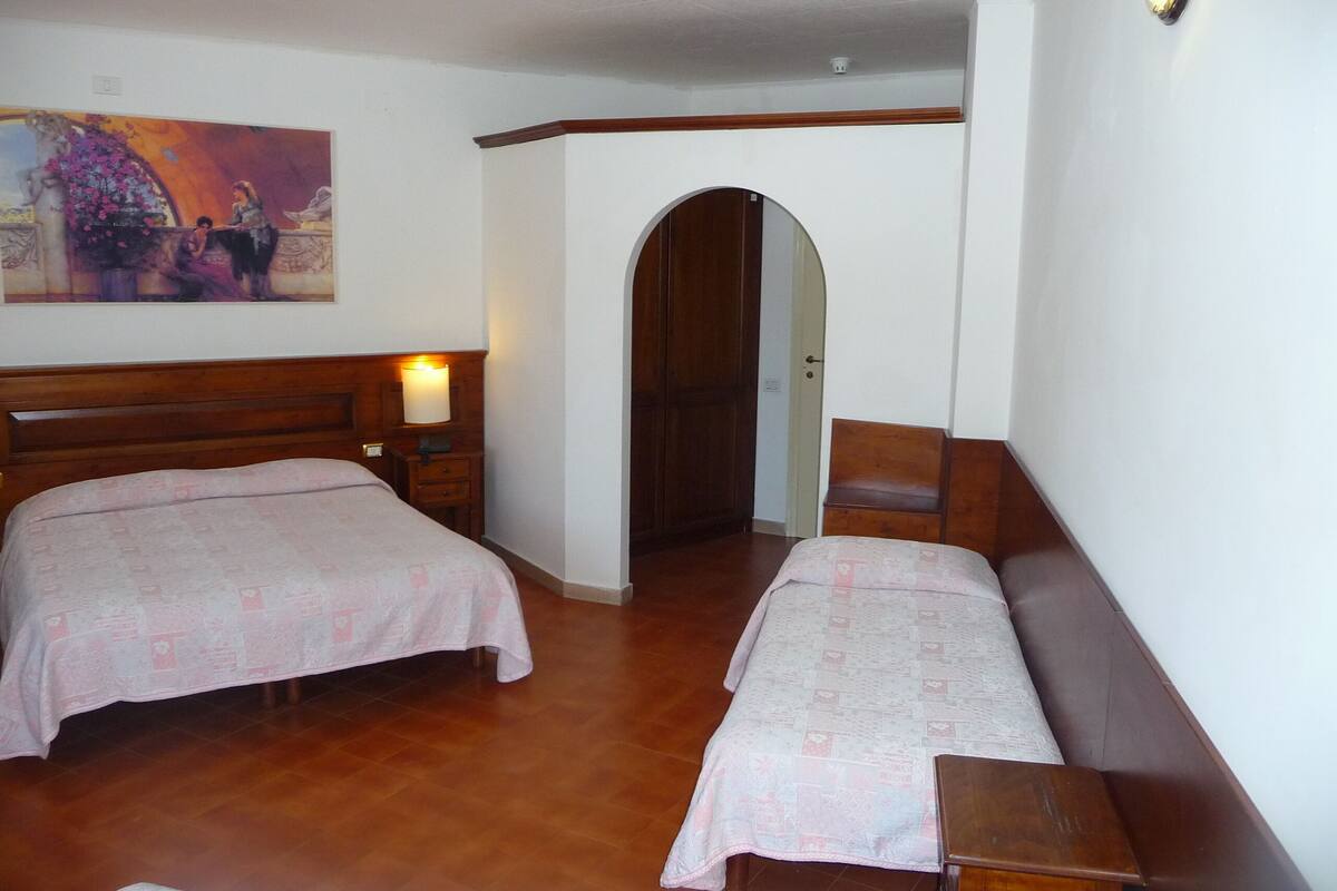 A hotel room with two beds, a wooden headboard, a nightstand with a lamp, a painting on the wall, and a door leading to another room.