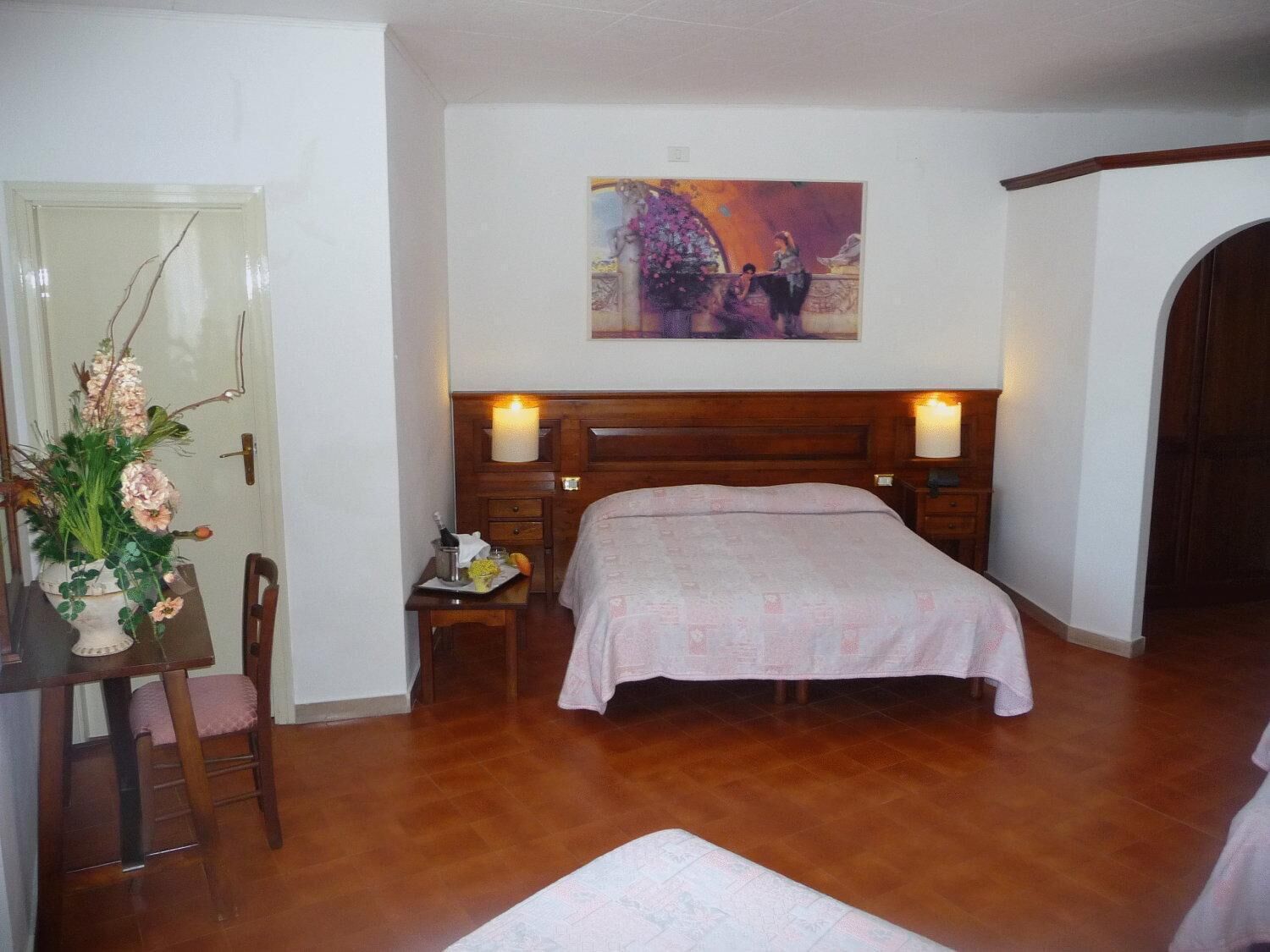 A bedroom with a wooden bed, a nightstand, a chair, a table with a vase of flowers, and a painting on the wall.