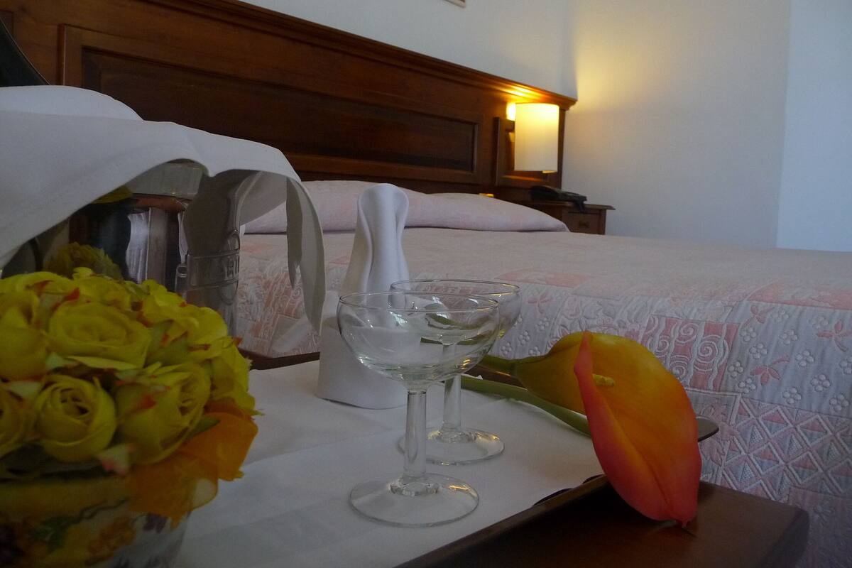 A neatly made bed with a patterned bedspread, a bedside table with a lamp, and a tray with a glass of water, a vase, and a bouquet of flowers.