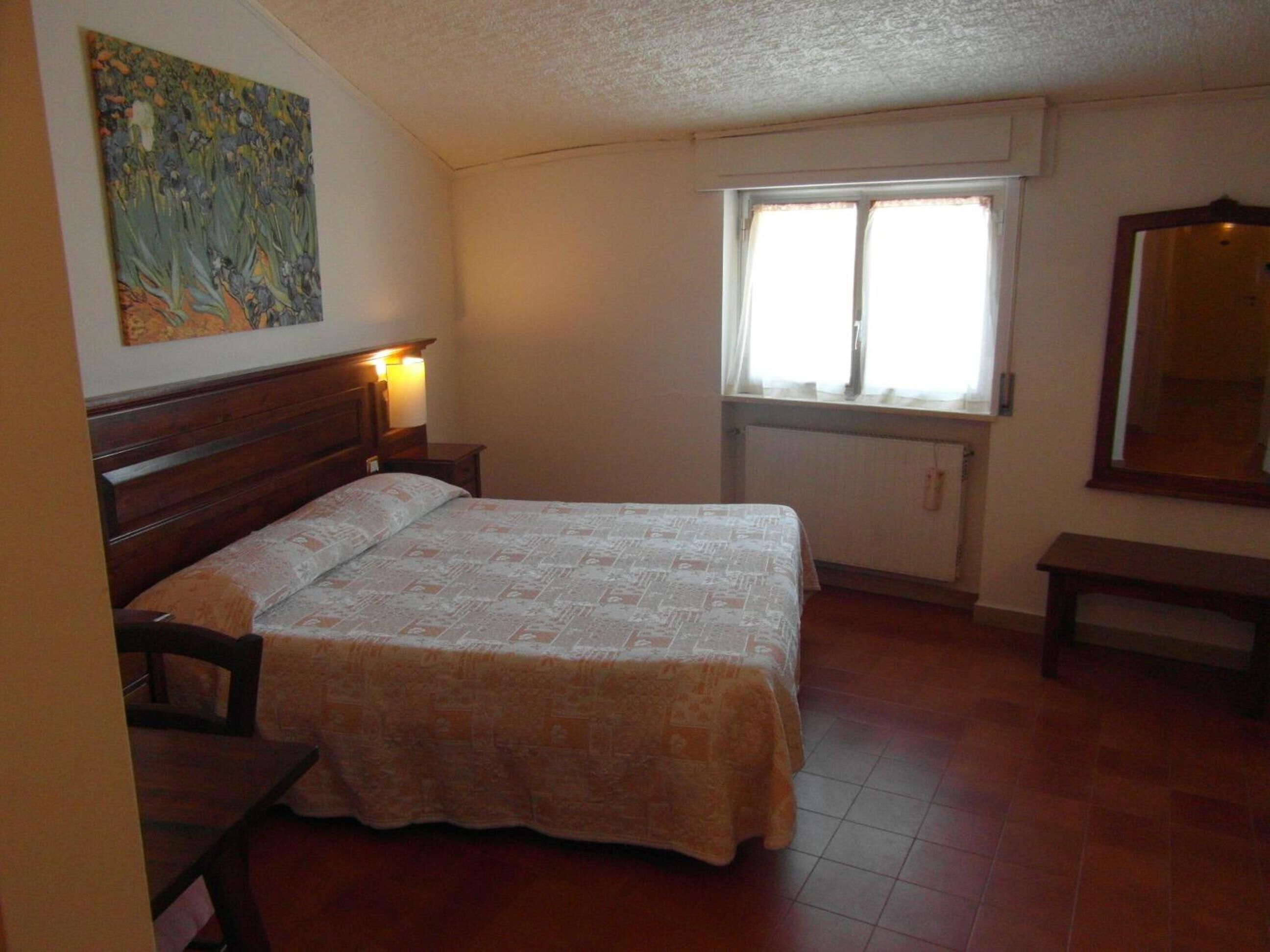 A bedroom with a bed, a nightstand, a bedside table, a window, and a painting on the wall.
