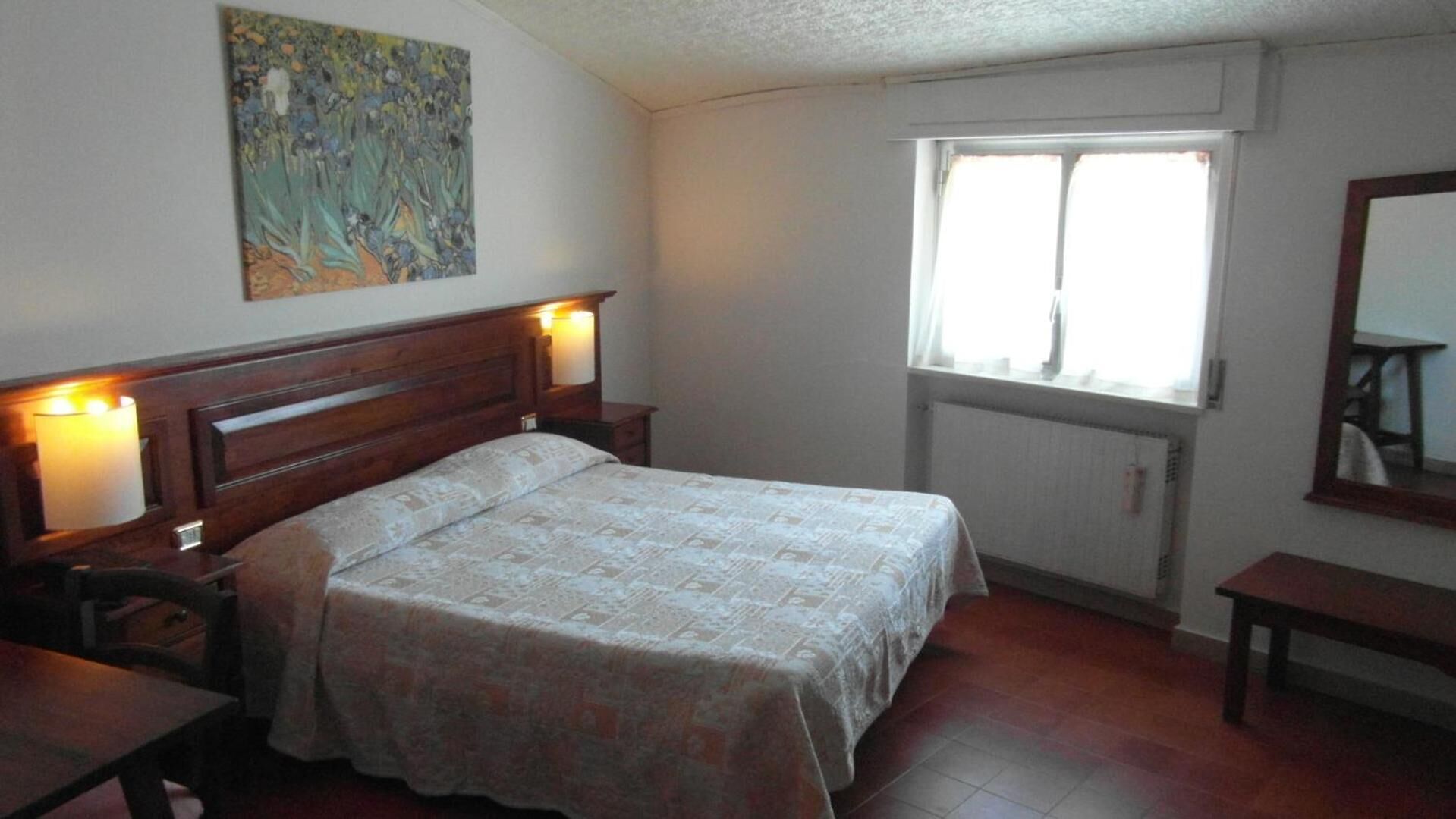 Una camera da letto con un letto, comodini, un tavolino, una finestra, un termosifone e un quadro appeso al muro.