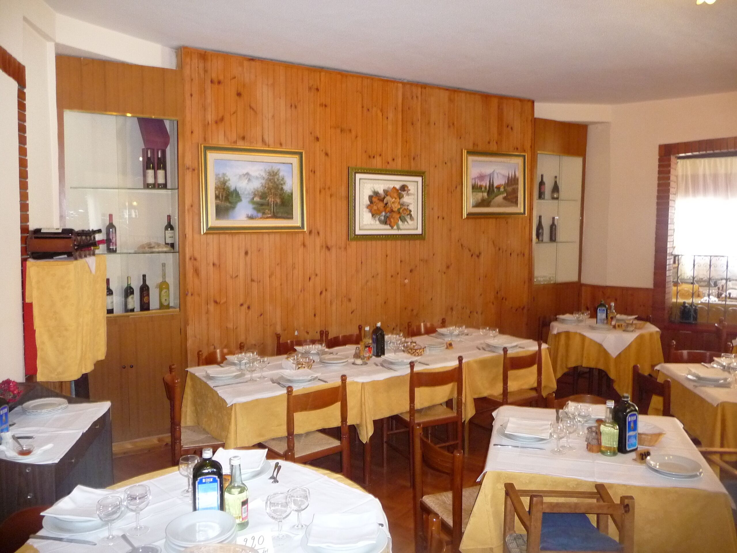 A dining area with wooden paneling, multiple tables set for dining, and framed landscape paintings on the walls.