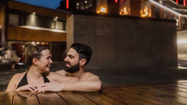 Un couple se trouve dans un espace extérieur avec une piscine chauffée, et on aperçoit un bâtiment moderne en arrière-plan.