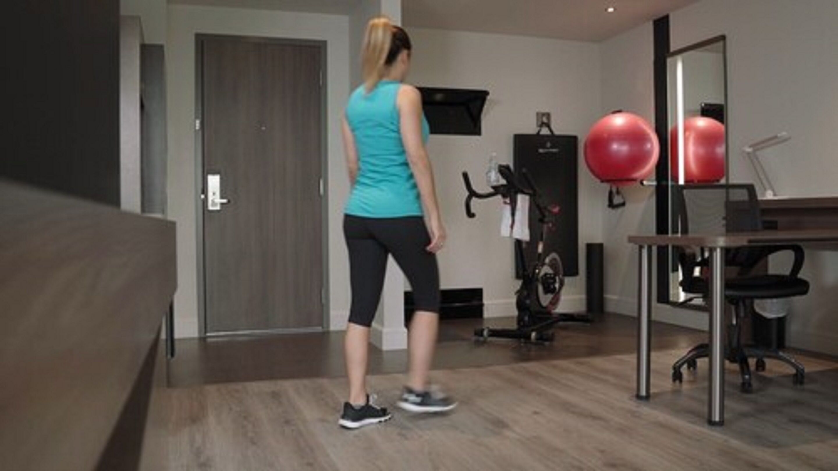 Habitación moderna con suelo de madera, escritorio, silla y equipo de ejercicio, incluyendo una bicicleta estática y pelotas de gimnasia rojas.