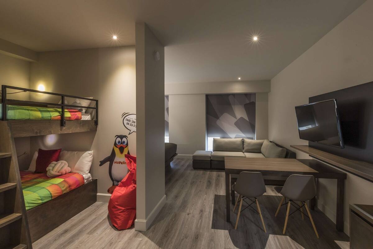 Un salon moderne avec un lit superposé, un fauteuil en forme de poire rouge, un canapé, une télévision et un coin repas.