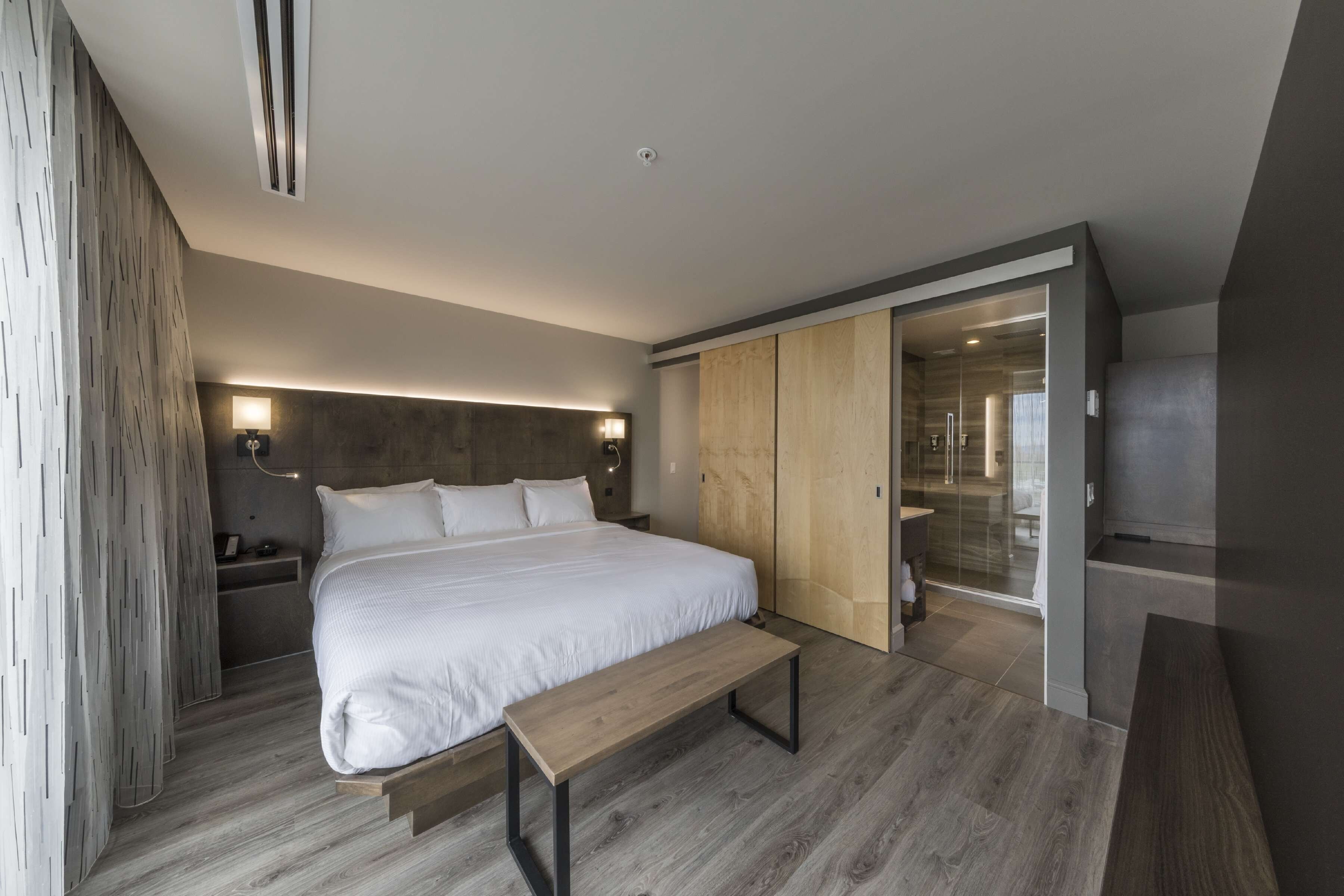 A modern bedroom with a large bed, wooden flooring, a bench, a wardrobe, and a bathroom visible through a glass door.