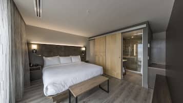 Une chambre moderne avec un grand lit, un plancher en bois, un banc, une armoire et une salle de bain visible à travers une porte en verre.