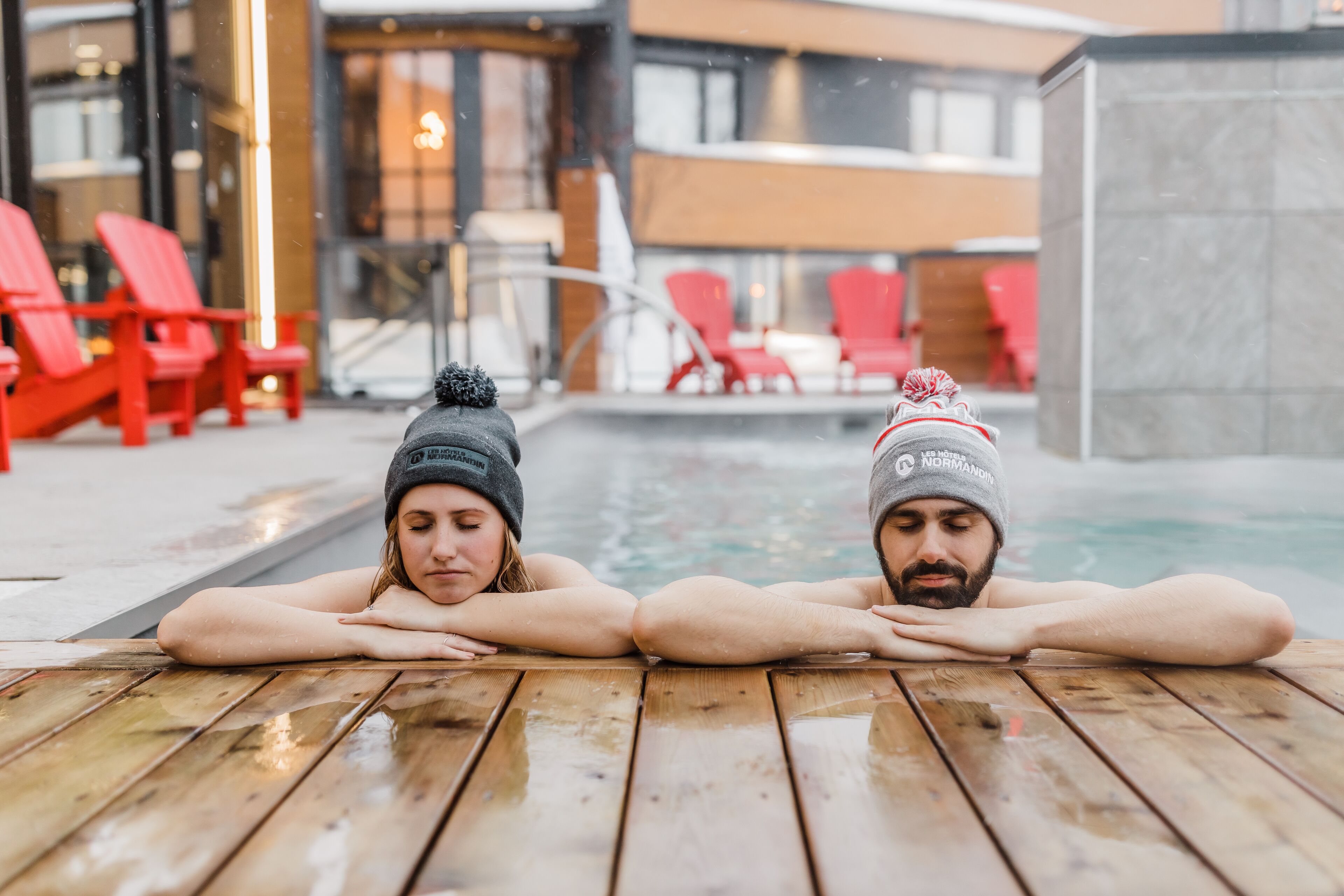 Dos personas descansando en un deck de madera junto a una piscina, con sillas rojas y un edificio moderno al fondo.