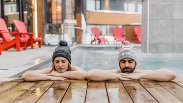 Dos personas descansando en un deck de madera junto a una piscina, con sillas rojas y un edificio moderno al fondo.