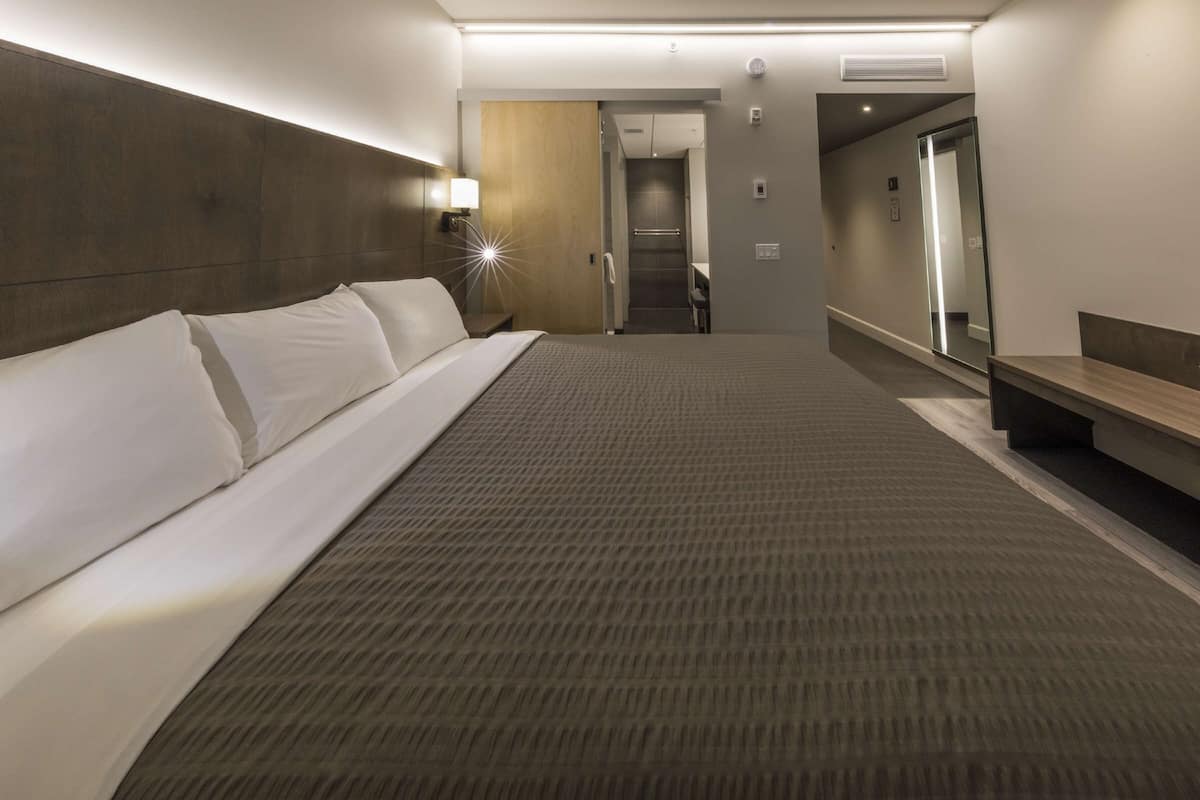 Une chambre d’hôtel avec un grand lit, une table de chevet, une commode, un miroir et, au fond, la porte de la salle de bain.