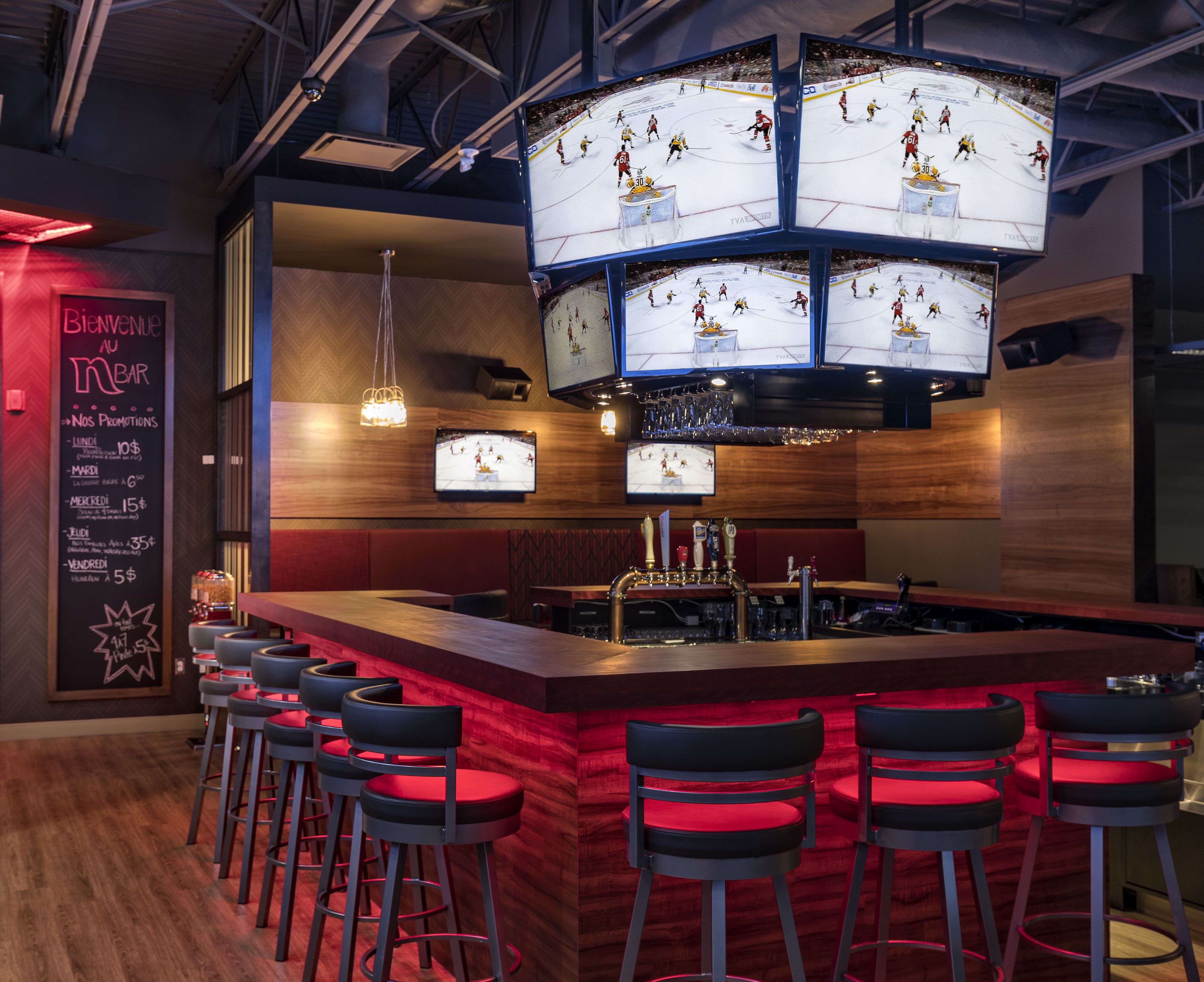 Un espace bar doté de plusieurs écrans diffusant des événements sportifs, d’un comptoir et de tabourets de bar rouges.