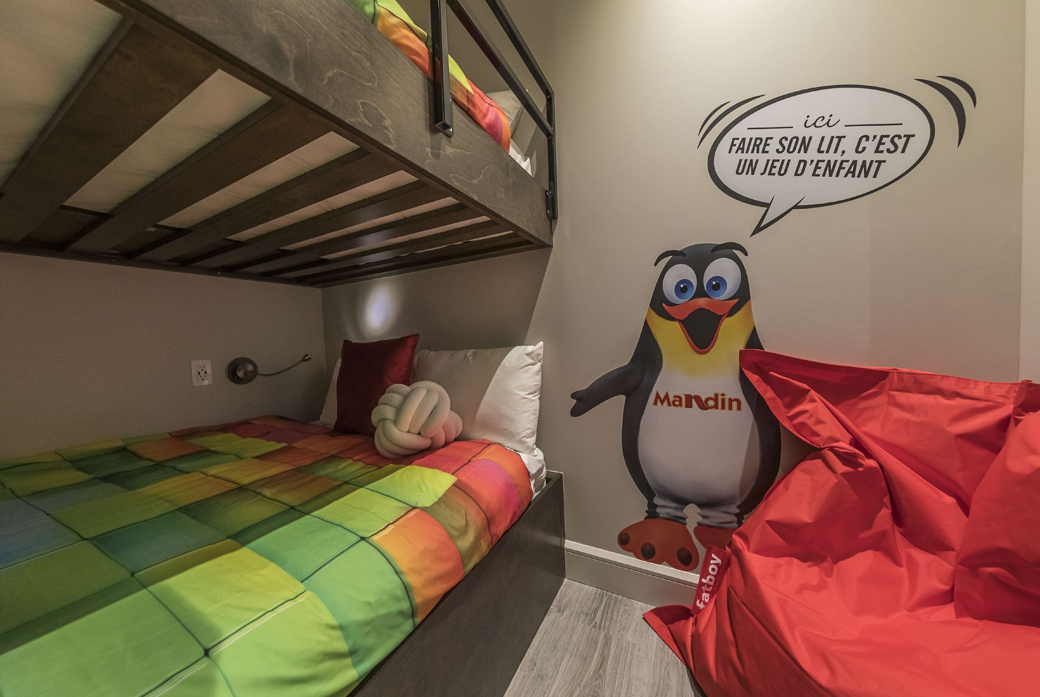 Una cama litera con colcha colorida, un peluche y un mural de un pingüino con burbuja de diálogo.