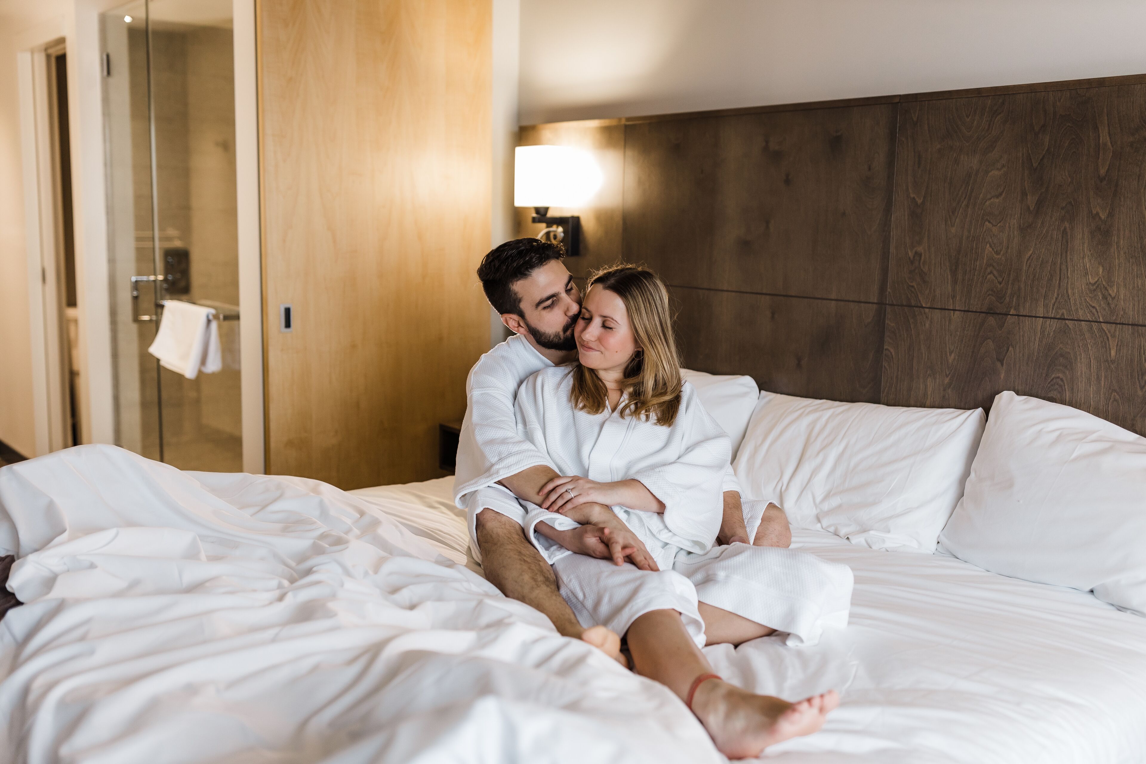 A couple in white robes sitting on a bed in a hotel room with a wooden headboard and a nightstand with a lamp.
