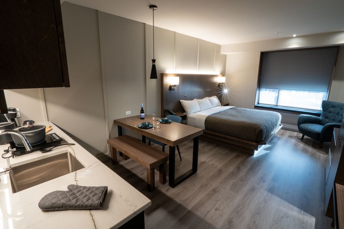 Une chambre d’hôtel compacte comprenant un coin cuisine, un lit avec une tête de lit, une table de chevet, un banc, une chaise et une grande fenêtre.