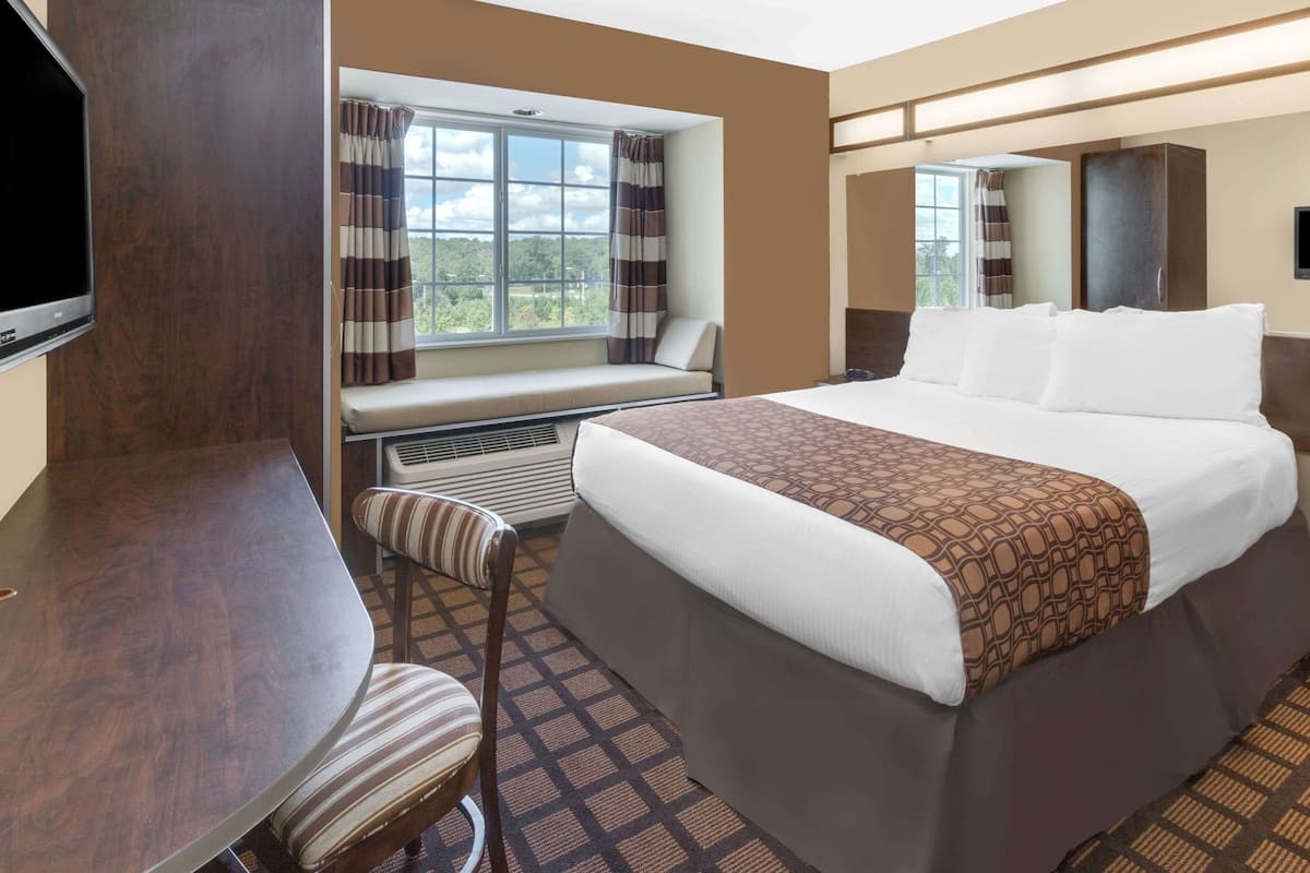 A hotel room with a bed, a desk, a chair, a window with curtains, and a television.