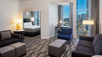 A hotel room with a bed, two armchairs, a desk, a large window with a city view, and a patterned carpet.