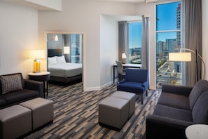 Ein Hotelzimmer mit einem Bett, zwei Sesseln, einem Schreibtisch, einem großen Fenster mit Blick auf die Stadt und einem gemusterten Teppich.