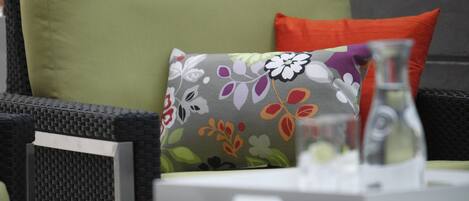 A wicker chair with a green cushion and a floral pillow, a small table with a glass pitcher, and a potted plant in the background.