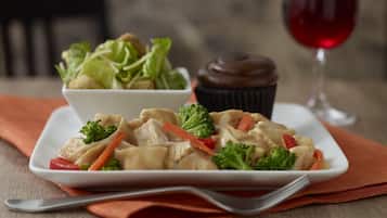 A plate of chicken with vegetables and a cupcake on a table with a wine glass in the background.