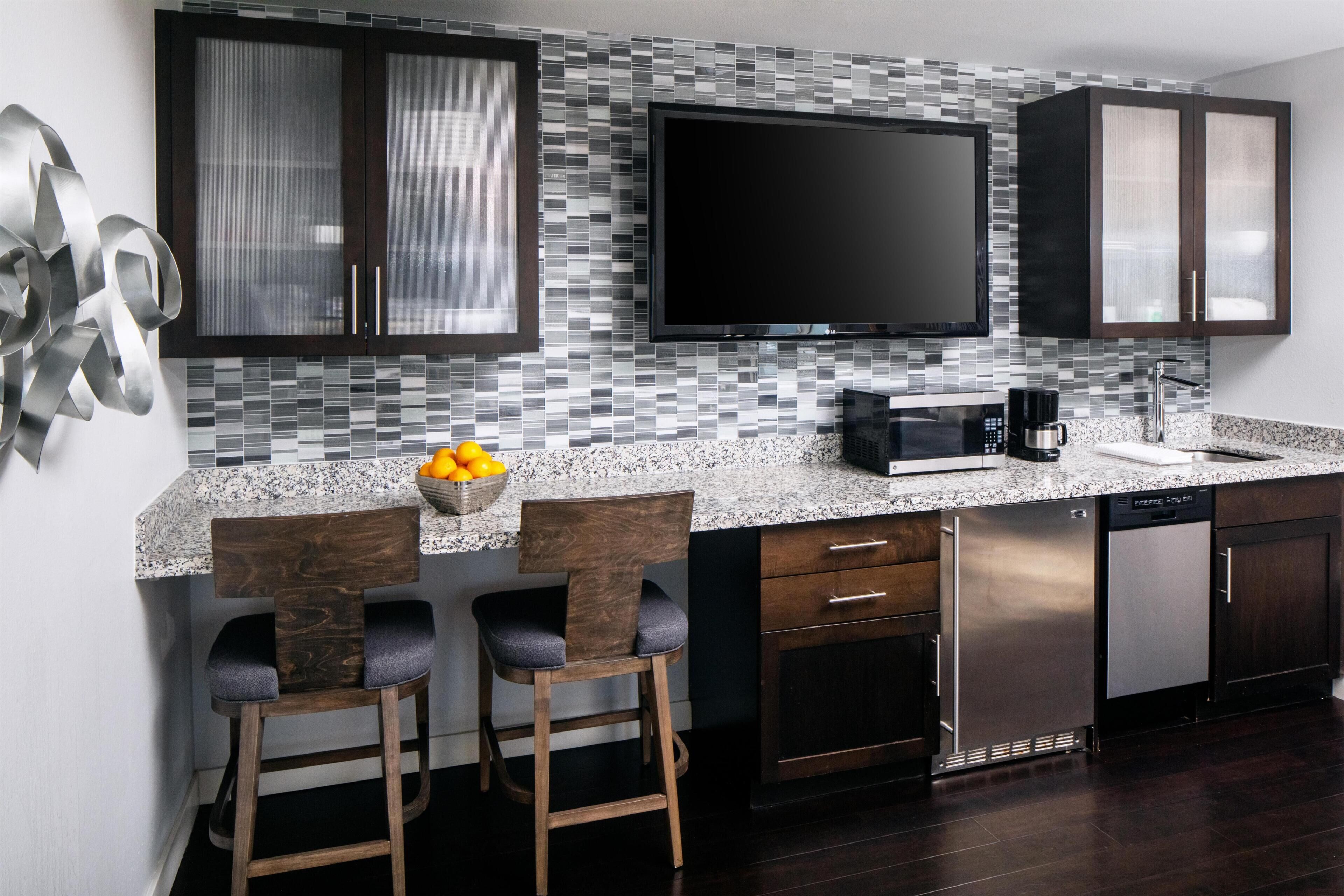A modern kitchen with a large flat-screen TV, dark cabinets, a granite countertop, and two bar stools.