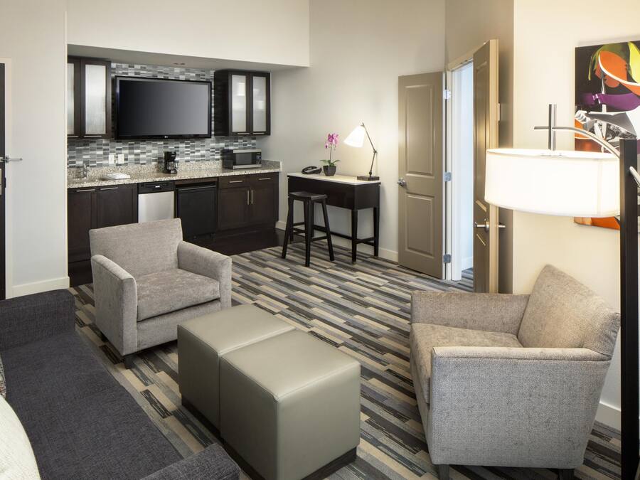 A hotel room with a kitchenette, two armchairs, a sofa, a coffee table, and a desk with a lamp.