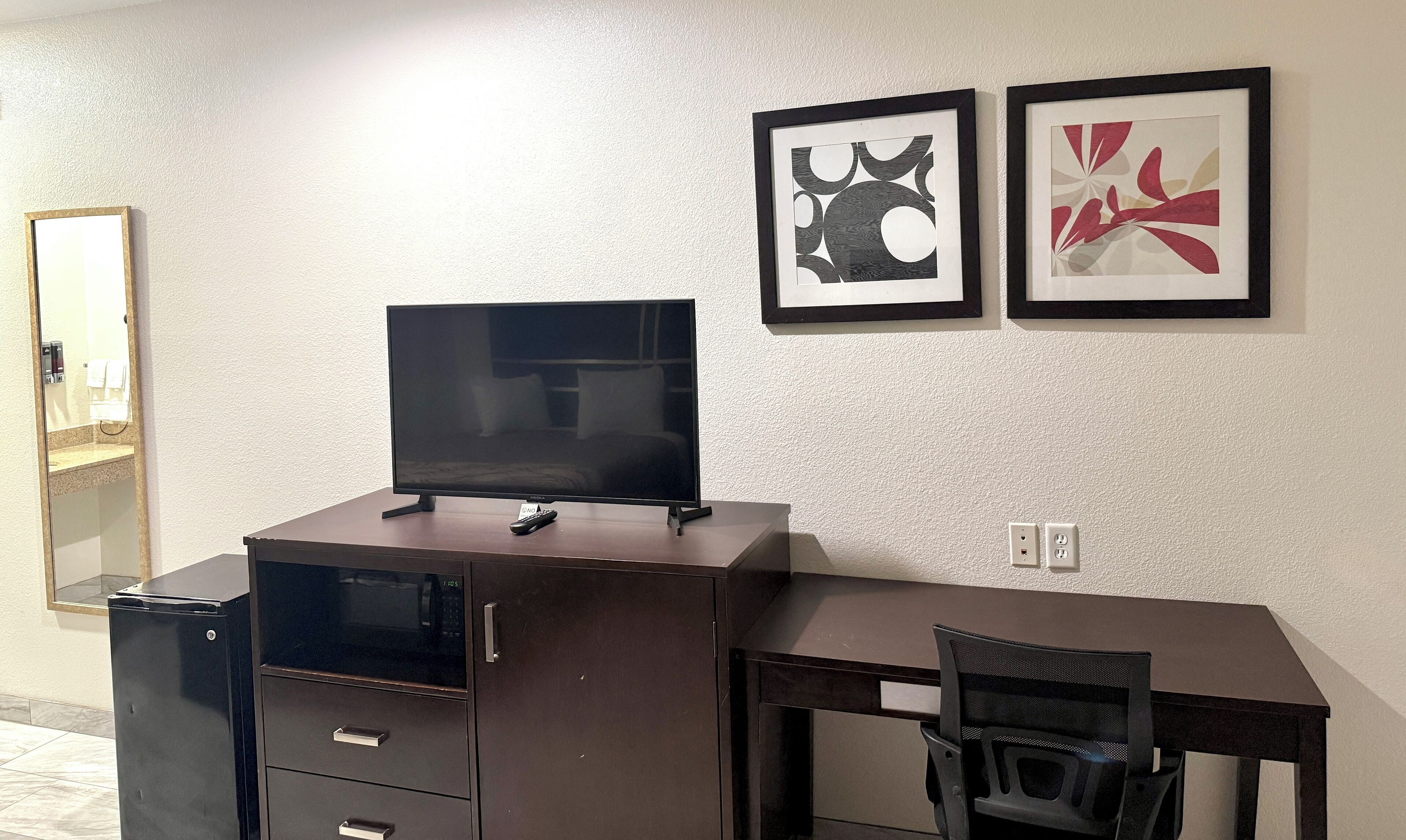 Une chambre d’hôtel compacte, dotée d’un bureau en bois foncé, d’un téléviseur à écran plat, d’un petit réfrigérateur et d’une chaise. Deux œuvres d’art abstraites encadrées ornent le mur.