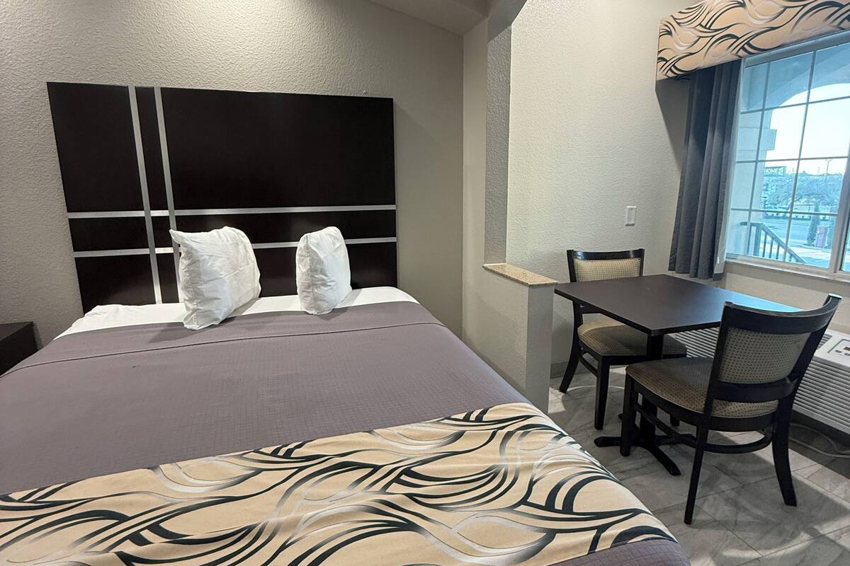 A hotel room with a large bed, a small table with chairs, a window with curtains, and a headboard with a light fixture.